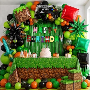 May include: A birthday party setup featuring a green pixelated tablecloth and matching square boxes. A tiered cake sits on the table. The backdrop includes a green fringe curtain, a banner reading HAPPY BIRTHDAY, and balloons shaped like a game controller, a sword, and stars in green, black, and red.