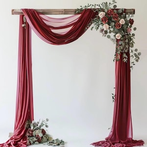 May include: A burgundy wedding arch with draped fabric and floral arrangements. The wooden frame is adorned with flowing burgundy fabric and accented with roses, greenery, and eucalyptus. The arch is set against a white backdrop.