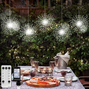 Può includere: Un set di cinque luci a stringa bianche a forma di fuochi d'artificio appese al soffitto sopra un tavolo apparecchiato per una cena. Le luci sono accese e illuminano il tavolo e l'area circostante. Il tavolo è apparecchiato con piatti, posate, bicchieri e una ciotola di cibo. C'è un telecomando per le luci sul tavolo.