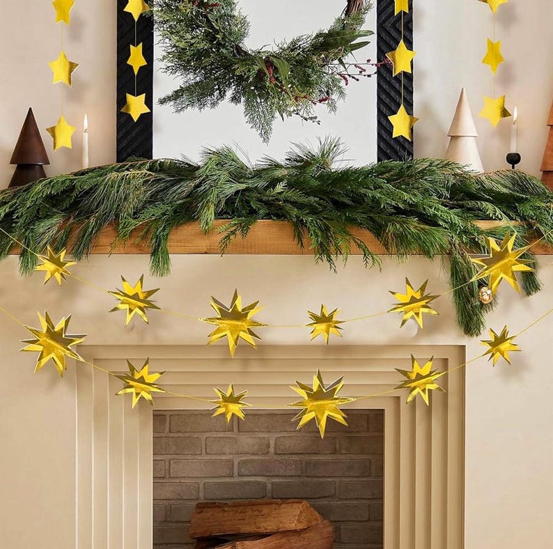 May include: A gold star garland with a string of paper stars hanging above a fireplace mantel. The garland is made of paper and has a metallic gold finish. The stars are all the same size and are evenly spaced apart. The garland is hanging from a wooden mantel and is decorated with a green garland of pine branches.