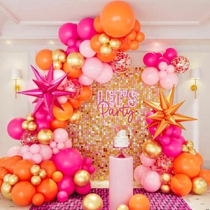 May include: A balloon arch made of pink, orange, and gold balloons with confetti balloons. The arch is decorated with two large star-shaped balloons. A cake sits on a table in front of the arch. The background is a gold sequin wall with the words "Let's Party" in neon pink letters.