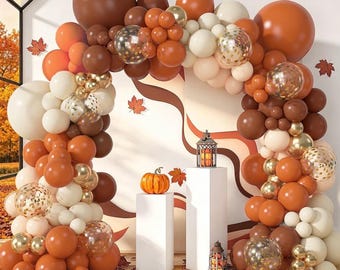 Puede incluir: Un gran arco de globos con una mezcla de globos en color marrón mate, naranja quemado, crema y dorado metálico, incluyendo globos transparentes con confeti dorado. Dos pedestales blancos sostienen una calabaza naranja y dos farolillos.