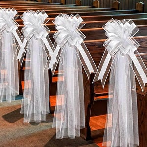 May include: White tulle bows with white satin ribbon tails attached to the back of church pews. The bows are tied with a knot and the tails are flowing down the back of the pews.