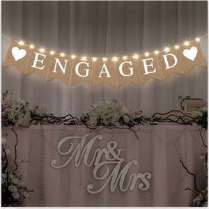 Puede incluir: Una pancarta de arpillera con letras blancas que deletrean "ENGAGED" y corazones blancos en cada extremo. La pancarta está adornada con luces blancas. Un letrero blanco "Mr. & Mrs." está sobre una mesa rosa con flores blancas.
