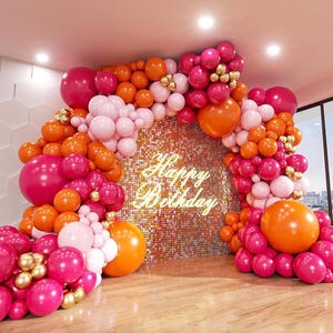Puede incluir: Un arco de globos rosa y naranja con detalles dorados, decorado con las palabras "Happy Birthday" en letras doradas. Los globos están dispuestos en cascada, creando un telón de fondo festivo para una fiesta.