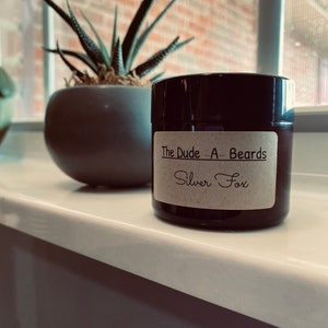 May include: A black jar with a brown label that reads "The Dude - A - Beards Silver Fox".