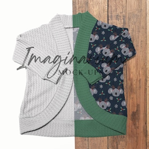 May include: A gray waffle-knit cardigan with an open front, displayed alongside a navy blue sleeve with a koala and holly print. The cardigan is laid out on a white surface and a wooden surface.