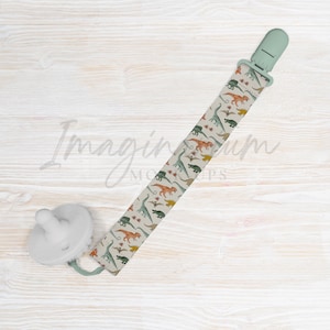 May include: A white pacifier clip with a green clip and a dinosaur pattern. The clip is on a white wooden surface.