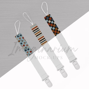 May include: Three baby pacifier clips with different patterned designs. One clip has a light blue background with orange and black jack-o'-lantern faces. The other two have striped and checkered patterns in neutral colors. Each clip has a white strap and a plastic clip.