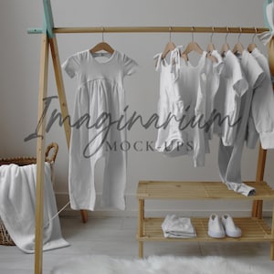 Op de afbeelding: Een houten kledingrek toont verschillende witte babykleding, waaronder een jumpsuit, jurken en een shirt met lange mouwen. Een opgevouwen deken ligt in een rieten mand en witte schoenen staan op een houten plank. De tekst "Imaginarium MOCK-UPS" is zichtbaar.