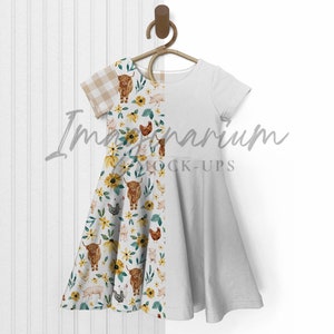 K&ouml;nnte beinhalten: Ein wei&szlig;es Kleid mit kurzen &Auml;rmeln und einem Farmtier-Druck auf der linken Seite. Das Kleid h&auml;ngt an einem Holzb&uuml;gel vor einem wei&szlig;en Hintergrund.