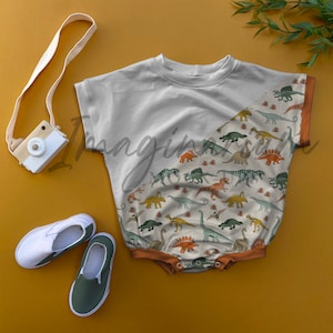 May include: A white and beige baby onesie with a dinosaur print. The onesie has a diagonal seam and a brown trim around the neckline and sleeves.