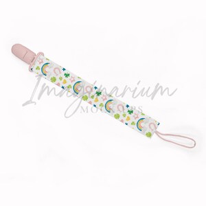 May include: A baby pacifier with a light pink nipple and a patterned strap. The strap features a white background with colorful rainbows, stars, horseshoes, and shamrocks. The pacifier is designed for infants.