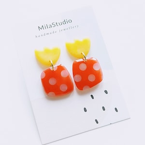 Puede incluir: Un par de pendientes hechos a mano con un diseño amarillo y rojo. Los pendientes tienen un semicírculo amarillo con un cuadrado rojo con lunares blancos que cuelga.