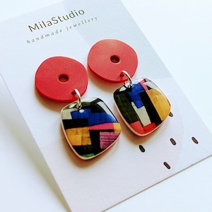 Op de afbeelding: Een paar rode en veelkleurige geometrische oorbellen met een geometrisch patroon. De oorbellen zijn handgemaakt en hebben een rode cirkel met een gat in het midden en een vierkant met een geometrisch patroon.