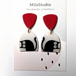 Puede incluir: Pendientes hechos a mano con una parte superior triangular roja y una base arqueada blanca con un diseño de gato negro. Los pendientes están conectados por un aro dorado. La tarjeta detrás de los pendientes dice "MilaStudio handmade jewellery."