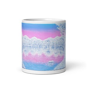 Puede incluir: Taza de cerámica blanca con una imagen pintada de una cordillera nevada reflejada en un lago al atardecer. El cielo es un degradado de rosa y azul.