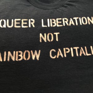 Peut inclure: T-shirt noir avec l'inscription "QUEER LIBERATION NOT RAINBOW CAPITALISM" imprimée en pochoir. Le texte est de couleur marron clair, contrastant avec le tissu sombre. Le t-shirt a un col rond.