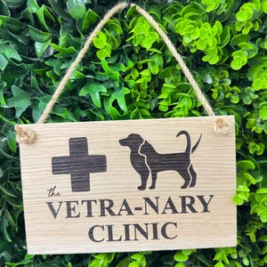 May include: Wooden sign for a veterinary clinic, featuring a black cross and a dog silhouette. The text "the VETRA-NARY CLINIC" is etched below. The sign hangs by a natural rope against a backdrop of green foliage.