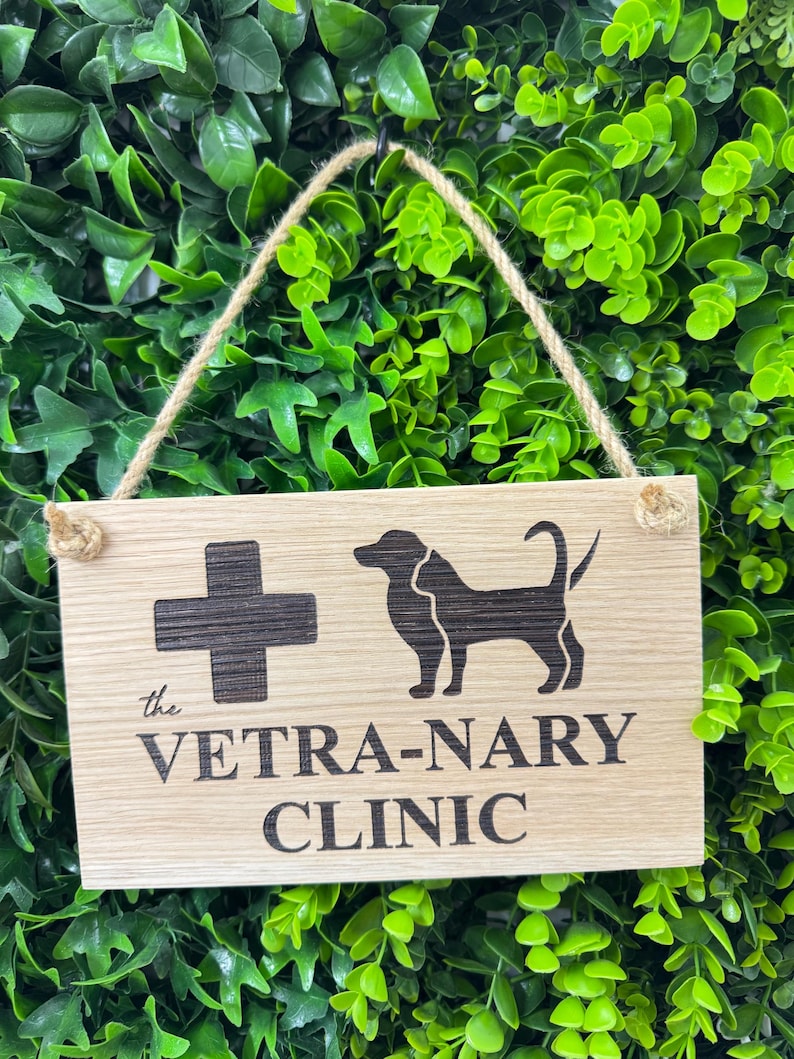 May include: Wooden sign for a veterinary clinic, featuring a black cross and a dog silhouette. The text "the VETRA-NARY CLINIC" is etched below. The sign hangs by a natural rope against a backdrop of green foliage.