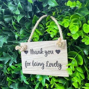 May include: A small, rectangular wooden sign with the words "Thank you for being Lovely" in black script, accented with heart symbols. The sign hangs from a natural rope against a backdrop of lush green foliage.