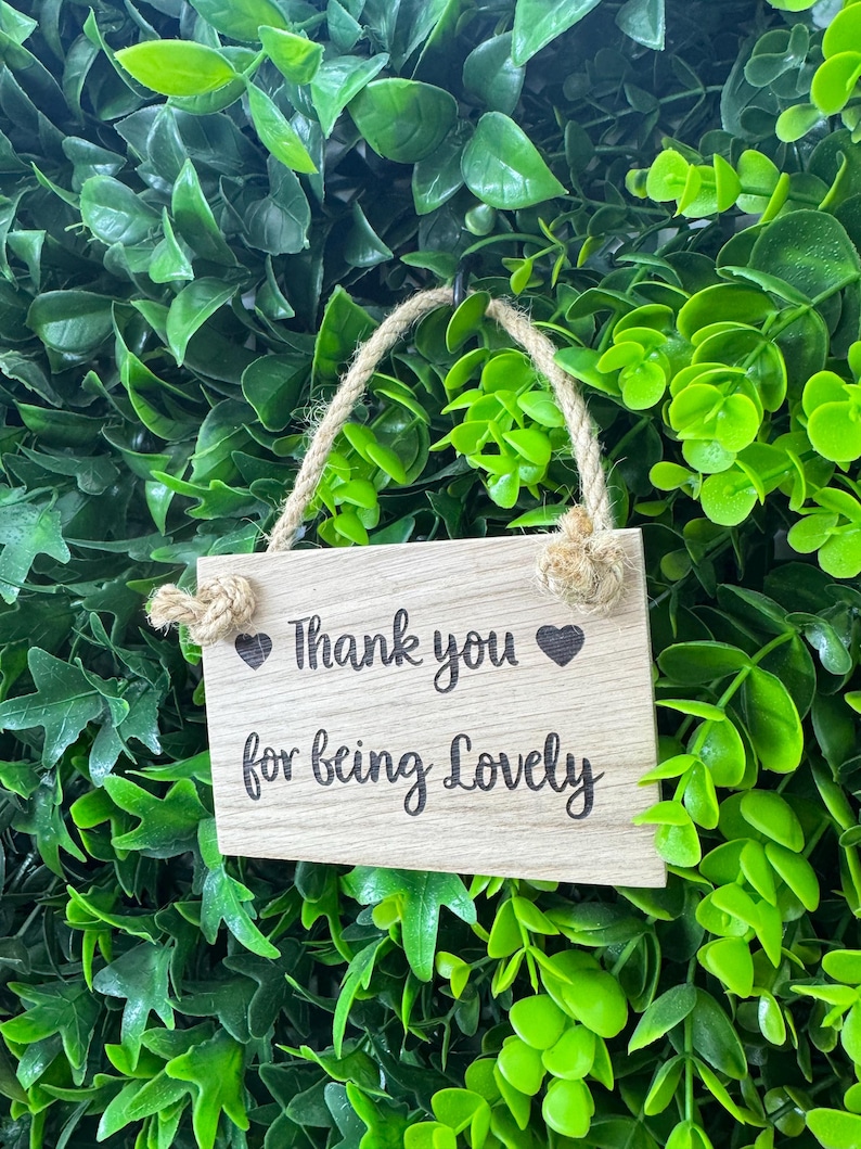 May include: A small, rectangular wooden sign with the words "Thank you for being Lovely" in black script, accented with heart symbols. The sign hangs from a natural rope against a backdrop of lush green foliage.