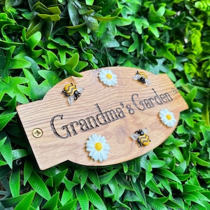 May include: Wooden garden sign with the text "Grandma's Garden", decorated with small white daisies and bee figurines. The sign is a light brown color and is mounted on a green leafy background.