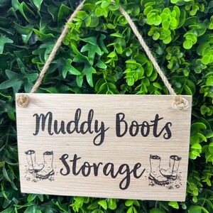May include: A rectangular wooden sign with the words "Muddy Boots Storage" in black script. The sign has a jute rope for hanging and features illustrations of boots. The background is a dense arrangement of green leaves.