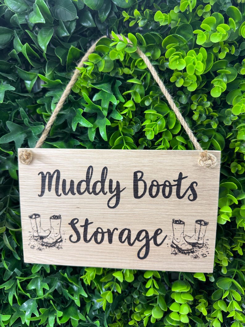 May include: A rectangular wooden sign with the words "Muddy Boots Storage" in black script. The sign has a jute rope for hanging and features illustrations of boots. The background is a dense arrangement of green leaves.