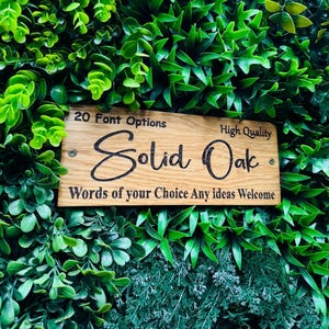 May include: A rectangular, light brown solid oak sign with black text. The sign reads "Solid Oak" in a cursive font, with additional text above and below. The sign is mounted on a background of green foliage.
