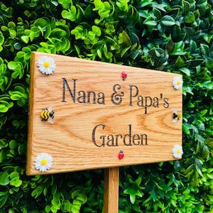 May include: Wooden garden sign with the text "Nana & Papa's Garden" in black lettering. The sign is decorated with small white daisies, red ladybugs, and yellow bumblebees.