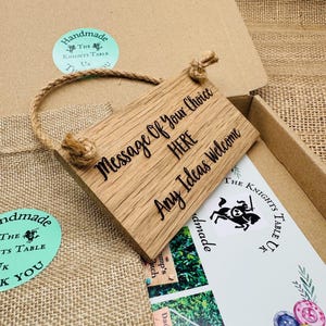May include: A rectangular wooden sign with the text "Message Of Your Choice HERE Any Ideas Welcome" in black script. The sign is attached to a natural rope and is inside a cardboard box. The box also contains a card with a knight logo.