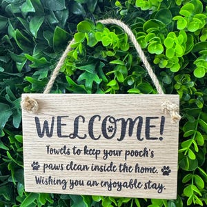May include: A rectangular wooden sign with the word "WELCOME!" in black lettering. The sign includes the text "Towels to keep your pooch's paws clean inside the home. Wishing you an enjoyable stay." A rope is attached for hanging.