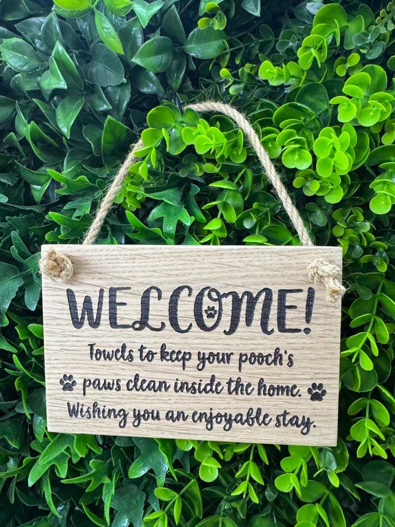 May include: A rectangular wooden sign with the word "WELCOME!" in black lettering. The sign includes the text "Towels to keep your pooch's paws clean inside the home. Wishing you an enjoyable stay." A rope is attached for hanging.