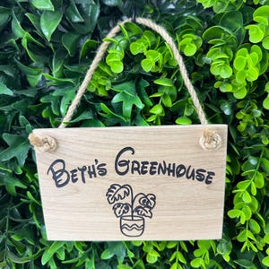 May include: A rectangular wooden sign with the text "Beth's Greenhouse" in black script, along with a potted plant illustration. The sign hangs from a jute rope against a backdrop of green foliage.