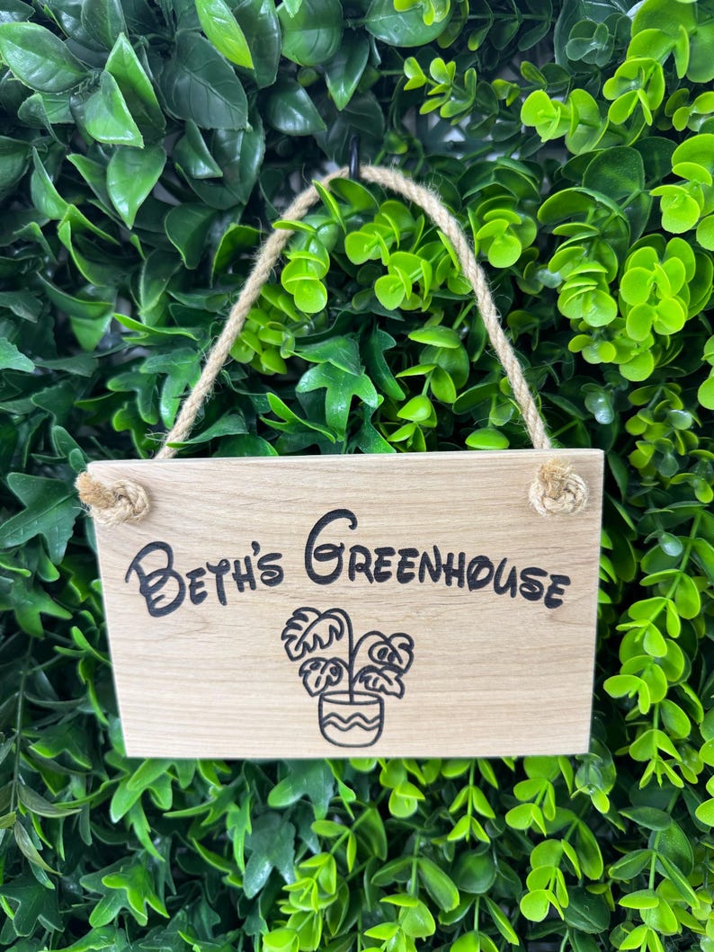 May include: A rectangular wooden sign with the text "Beth's Greenhouse" in black script, along with a potted plant illustration. The sign hangs from a jute rope against a backdrop of green foliage.