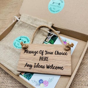 May include: A wooden sign with the text "Message Of Your Choice HERE Any Ideas Welcome" hangs in a cardboard box. A small burlap bag and a card with button illustrations are also in the box. The sign has a rope hanger.
