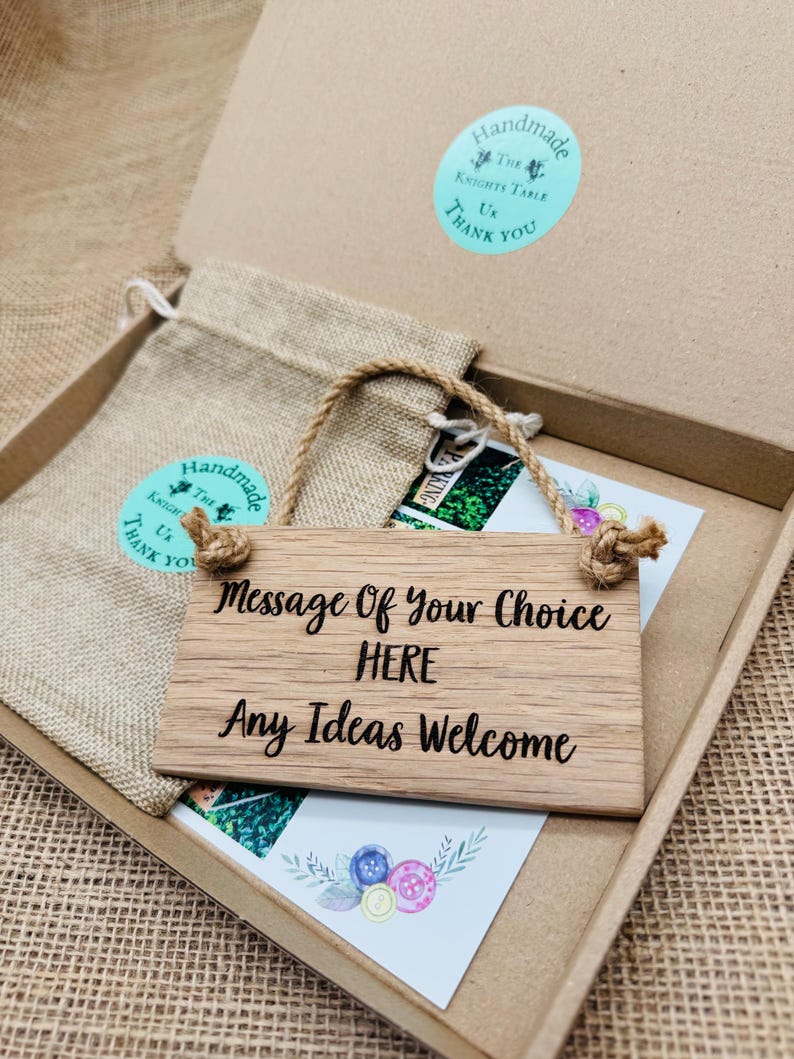 May include: A wooden sign with the text "Message Of Your Choice HERE Any Ideas Welcome" hangs in a cardboard box. A small burlap bag and a card with button illustrations are also in the box. The sign has a rope hanger.