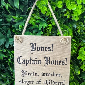 May include: A wooden sign with the text "Bones! Captain Bones! Pirate, wrecker, slayer of children!" in a gothic font. The sign is rectangular, with a natural wood grain finish, and is suspended by a rope. The background is a wall of green foliage.
