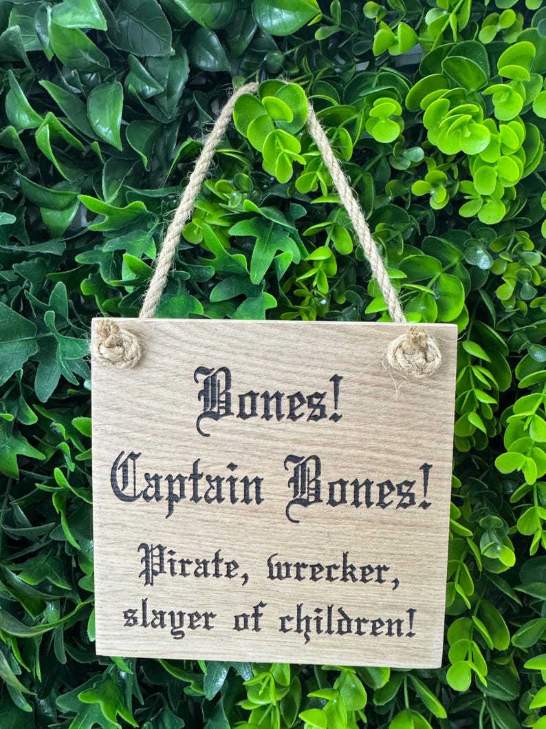 May include: A wooden sign with the text "Bones! Captain Bones! Pirate, wrecker, slayer of children!" in a gothic font. The sign is rectangular, with a natural wood grain finish, and is suspended by a rope. The background is a wall of green foliage.