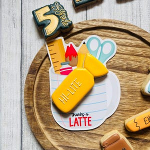 May include: A collection of school-themed cookies including a yellow highlighter cookie labeled HI-LITE, a mug-shaped cookie with the text thanks a LATTE, and dark green number five cookies with ruler details. Additional cookies include a lunch bag shape.