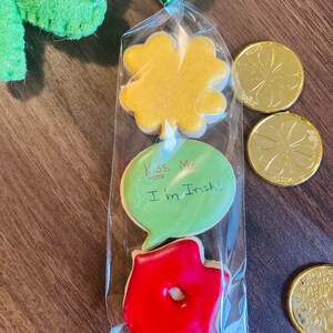 Può includere: Un sacchetto di plastica trasparente riempito con tre biscotti decorati. I biscotti hanno la forma di un quadrifoglio, una bolla di dialogo e una labbra rosse. Il biscotto a forma di bolla di dialogo ha la scritta "Kiss Me I'm Irish!" Il sacchetto è legato con un nastro verde e bianco.
