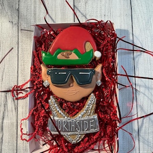May include: A box of decorated sugar cookies. One cookie is an elf hat with a green brim and a white pom-pom. Another cookie is a face wearing sunglasses and earrings. The third cookie has a silver necklace with the word "NORTHSIDE".