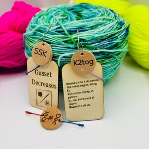 May include: Wooden knitting stitch markers with text, including "SSK" and "K2tog", hang in front of colorful yarn. The markers have instructions for knitting techniques. The background features bright pink, green, and yellow yarn.