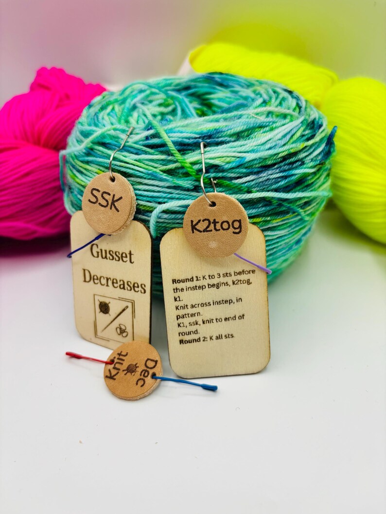 May include: Wooden knitting stitch markers with text, including "SSK" and "K2tog", hang in front of colorful yarn. The markers have instructions for knitting techniques. The background features bright pink, green, and yellow yarn.
