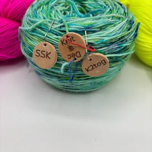 May include: A ball of teal, green, and blue yarn with three small wooden tags attached. The tags have the text "SSK", "Knit Dec", and "K2tog" printed on them. Two other balls of yarn in bright pink and neon yellow are in the background.