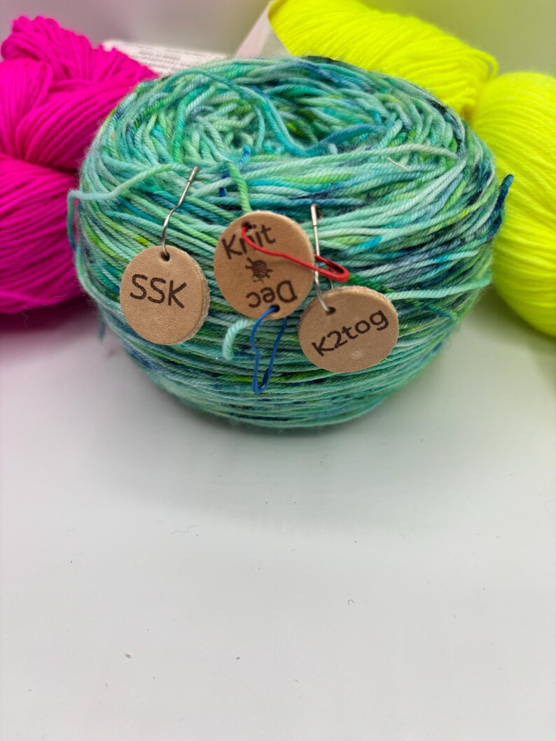 May include: A ball of teal, green, and blue yarn with three small wooden tags attached. The tags have the text "SSK", "Knit Dec", and "K2tog" printed on them. Two other balls of yarn in bright pink and neon yellow are in the background.