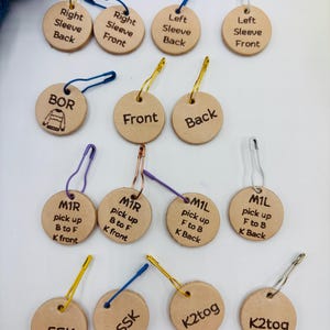 May include: A selection of circular, light brown leather knitting stitch markers. Each marker features text such as "Left Sleeve Front" and "K2tog", with a safety pin and a coloured string or wire loop. Ideal for knitters.