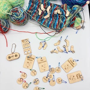 May include: A collection of wooden knitting notions including stitch markers, row counters, and progress keepers shaped like socks and circles. The items feature engraved text such as SSK, K2Tog, Toe Decreases, and Gusset Decreases, accompanied by colourful metal stitch marker rings, displayed alongside several balls of multi-coloured yarn and a partially knitted sock.