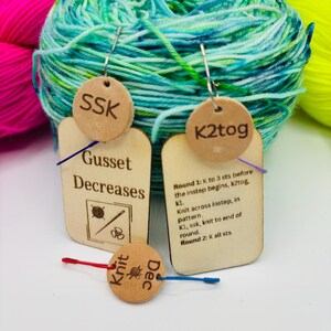 May include: Wooden knitting stitch markers with text including "SSK", "K2tog", "Gusset Decreases", "Knit Dec", and knitting instructions. The markers are attached to a blue and green yarn ball, with pink and yellow yarn in the background.